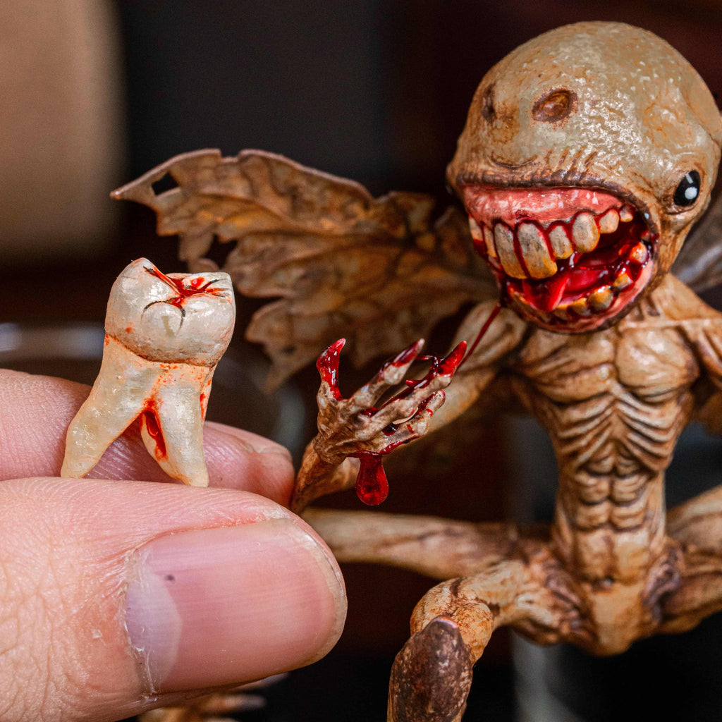 3D printed Tooth Fairy Hellboy figurine with skeletal wings, sharp claws, and a grotesque grin holding a tooth, surrounded by scattered white teeth. Hand-painted in bone, red, and brown tones, this dark fantasy collectible doubles as a tooth holder, making it perfect for gothic decor, horror props, or a unique keepsake for children’s lost teeth.