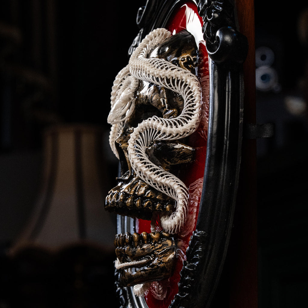 real snake skeleton is artfully curled around a 3D printed human skull, mounted on a deep red velvet background inside an ornate Gothic-style frame. The crimson backdrop intensifies the visual contrast, evoking a sense of blood, passion, and the eternal mystery of life and death.