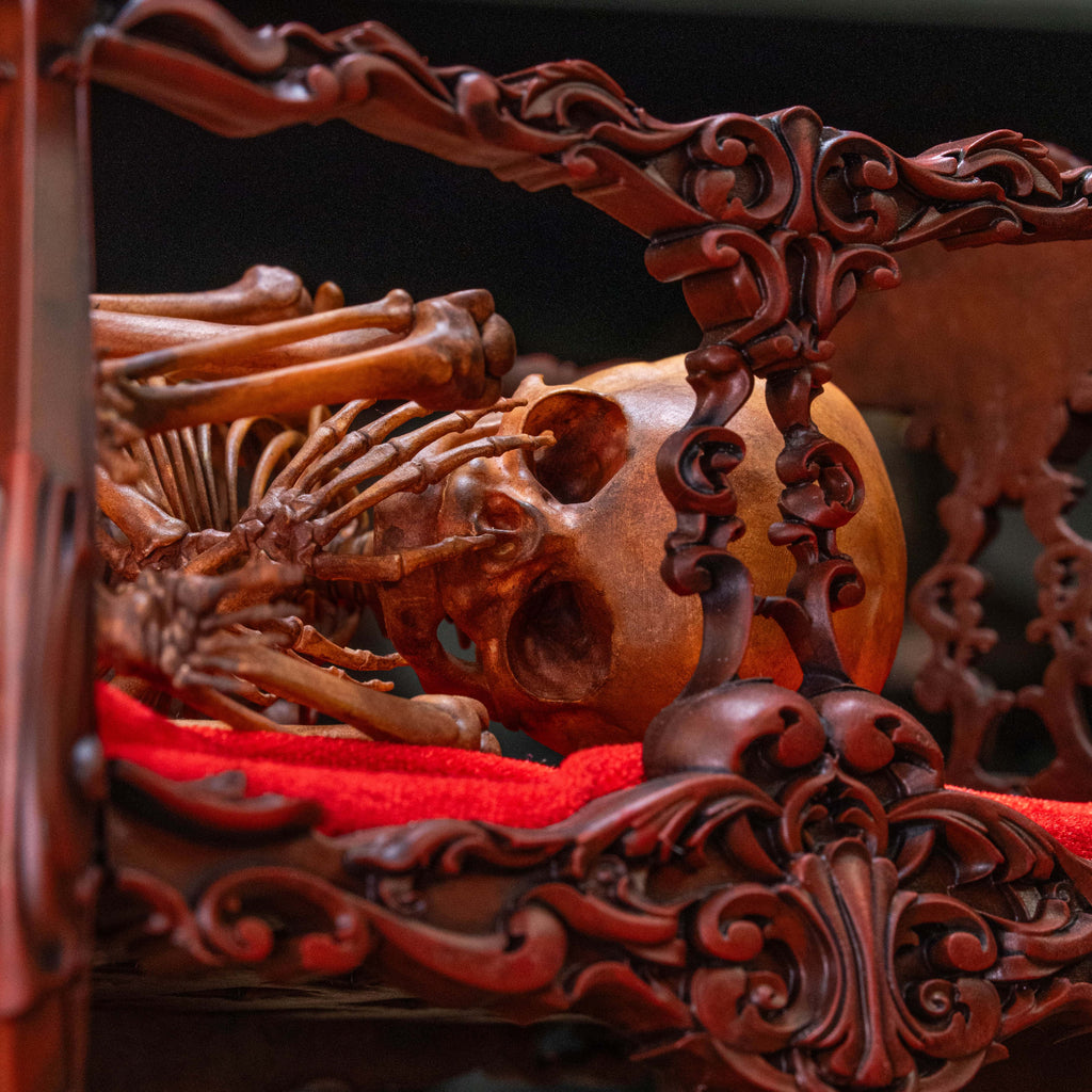 May include: A human skull and skeletal remains lying on a red velvet cushion inside a carved wooden frame. The frame is intricately detailed with ornate carvings and a dark red finish.